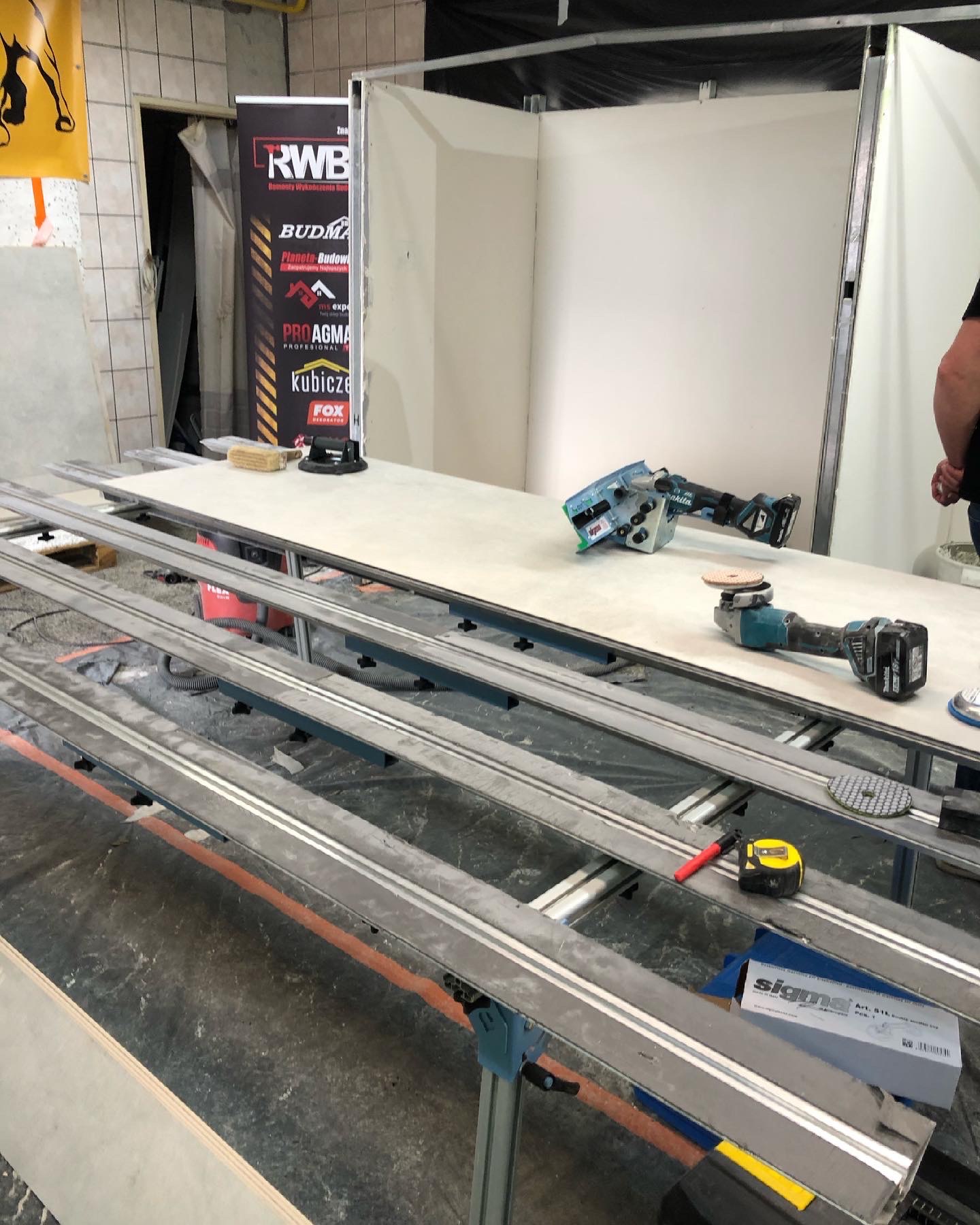 Przygotowane narzędzia, w tym szlifierki i przymiary, na stole warsztatowym w trakcie prac wykończeniowych, z widocznym stoiskiem reklamowym w tle.