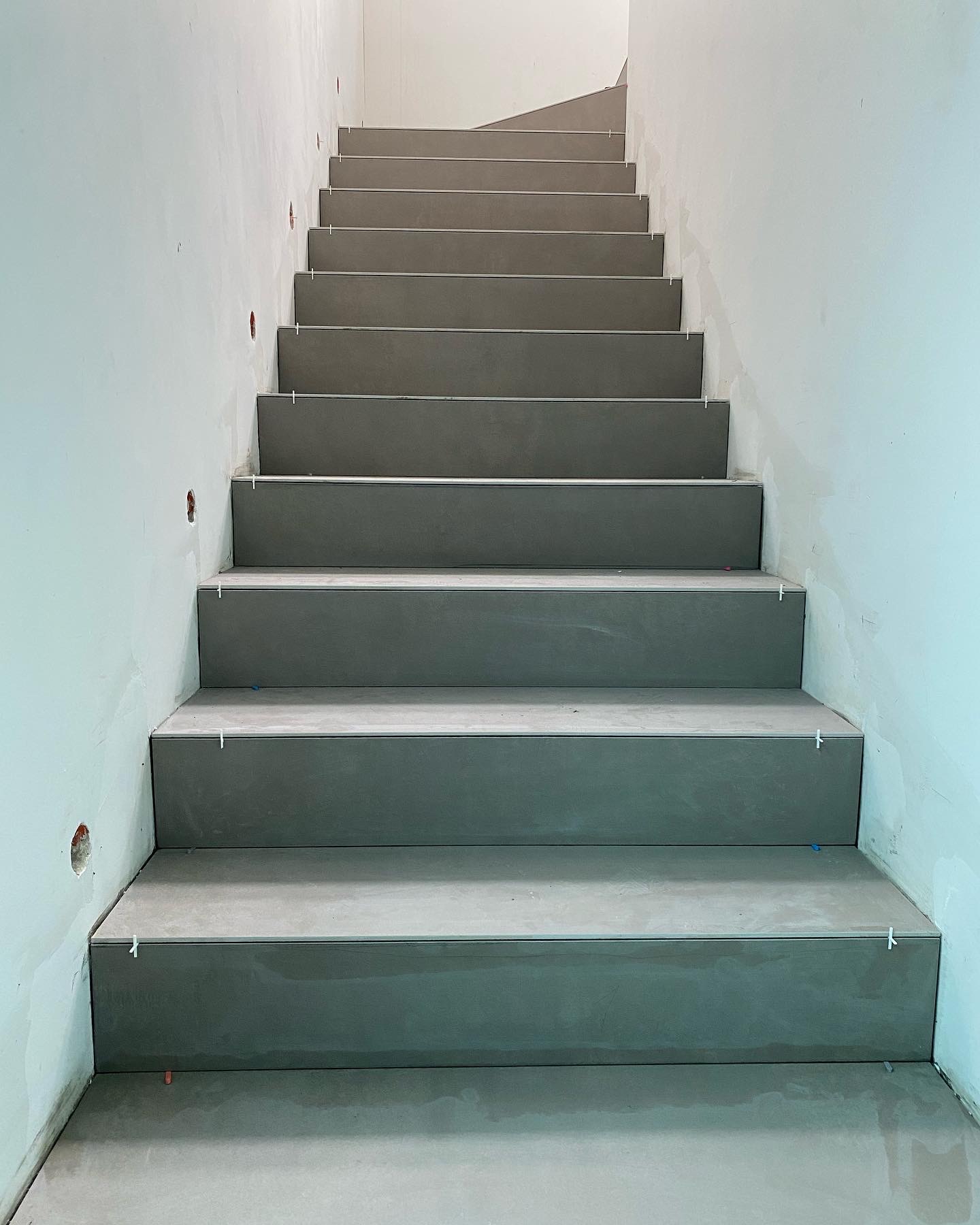 Schody wyłożone szarymi płytkami w trakcie montażu, widoczne krzyżyki dystansowe, ściany częściowo przygotowane do malowania z widocznymi otworami na gniazdka.