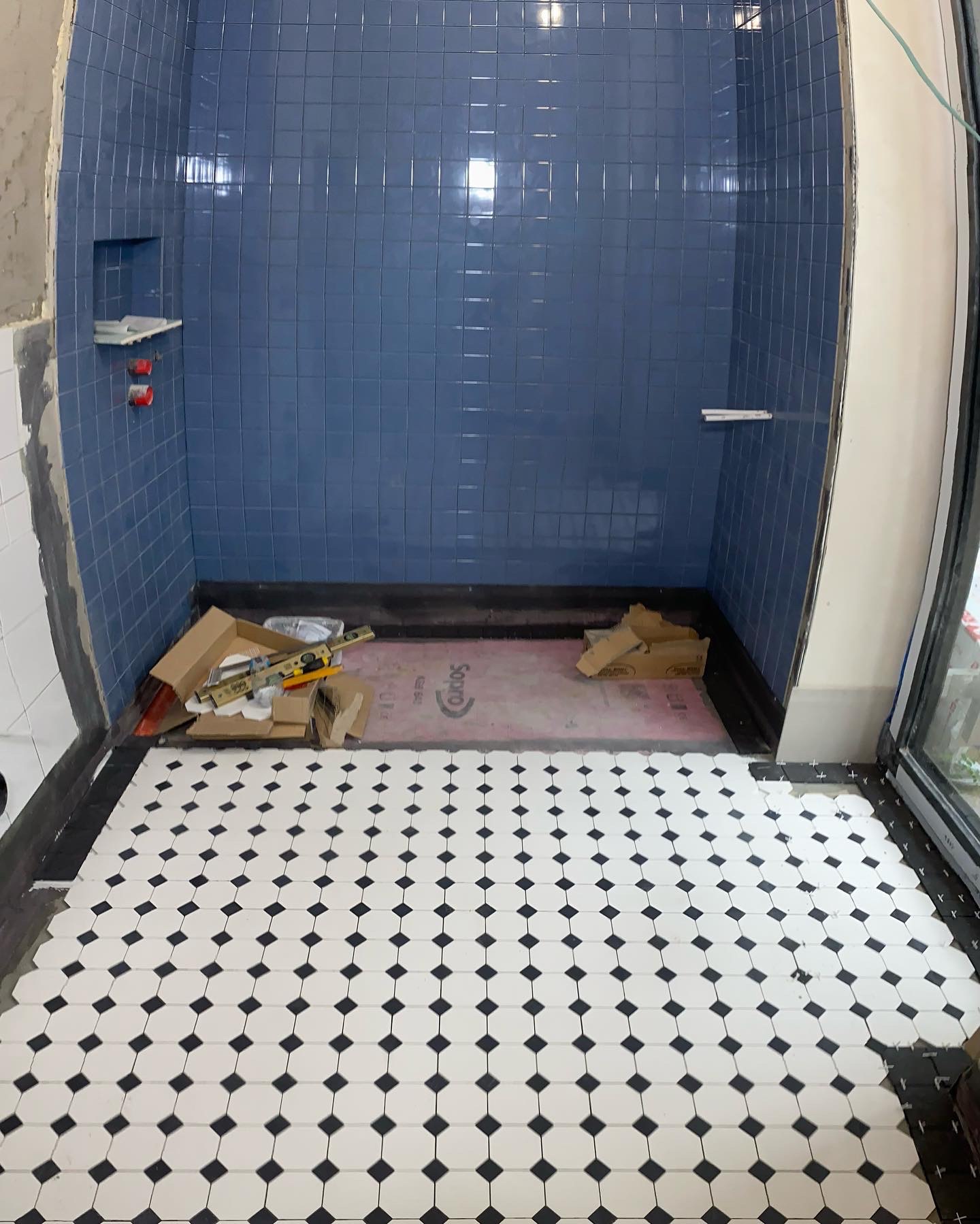 Niebieska glazura ścienna w kabinie prysznicowej z wnęką, biało-czarna mozaika podłogowa w trakcie układania, widoczne narzędzia i kartony.
