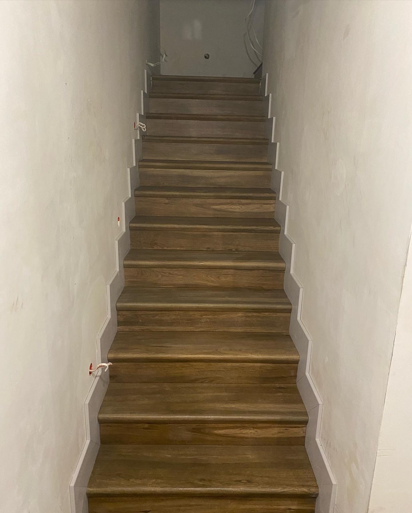 Drewniane schody z szarą listwą przypodłogową na tle surowej ściany, widoczne wystające przewody elektryczne.
