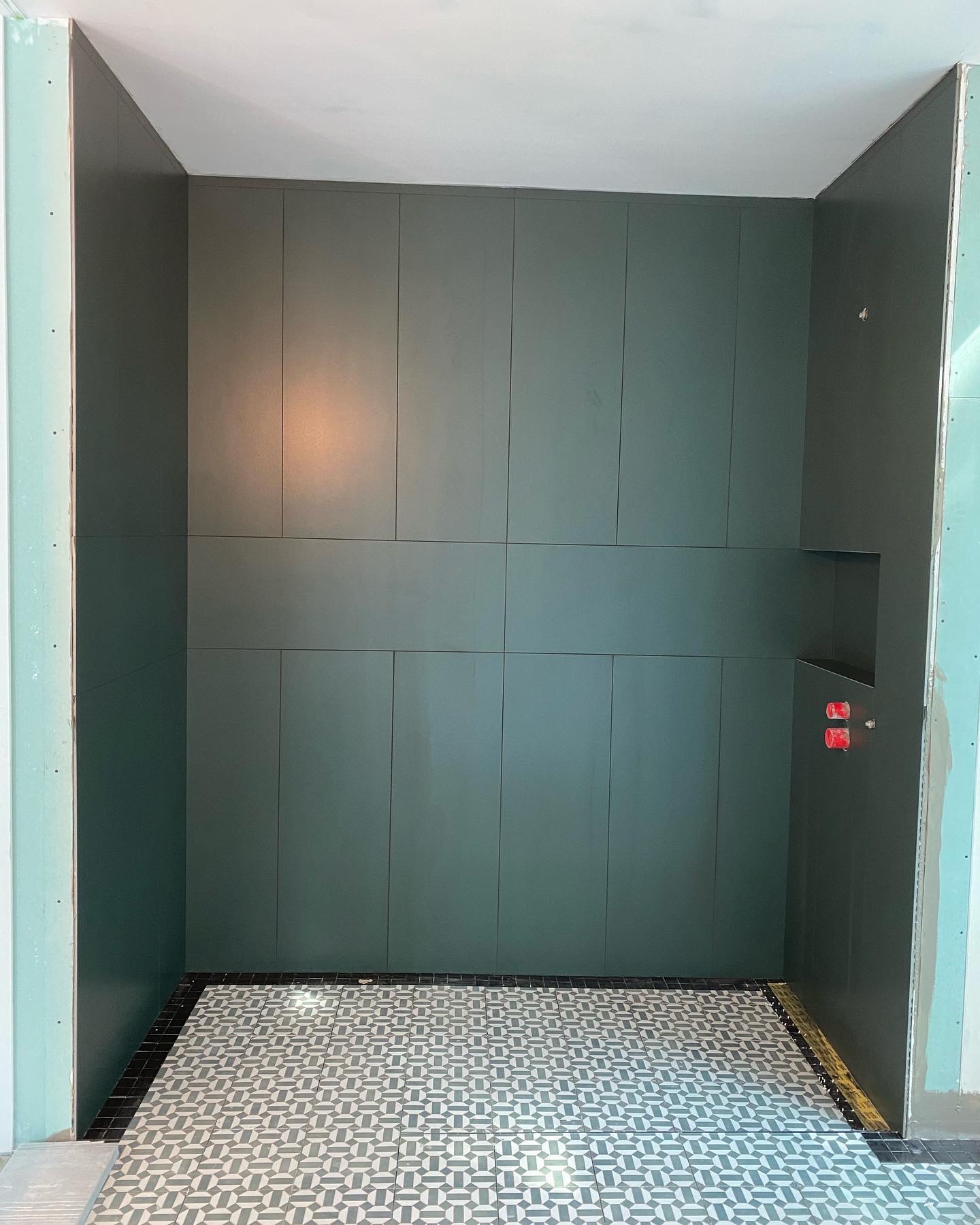 Wnętrze łazienki w trakcie prac wykończeniowych: ściany pokryte ciemnozielonymi panelami z wnęką, podłoga wyłożona wzorzystymi płytkami w odcieniach szarości i czerni, widoczne fragmenty surowych...