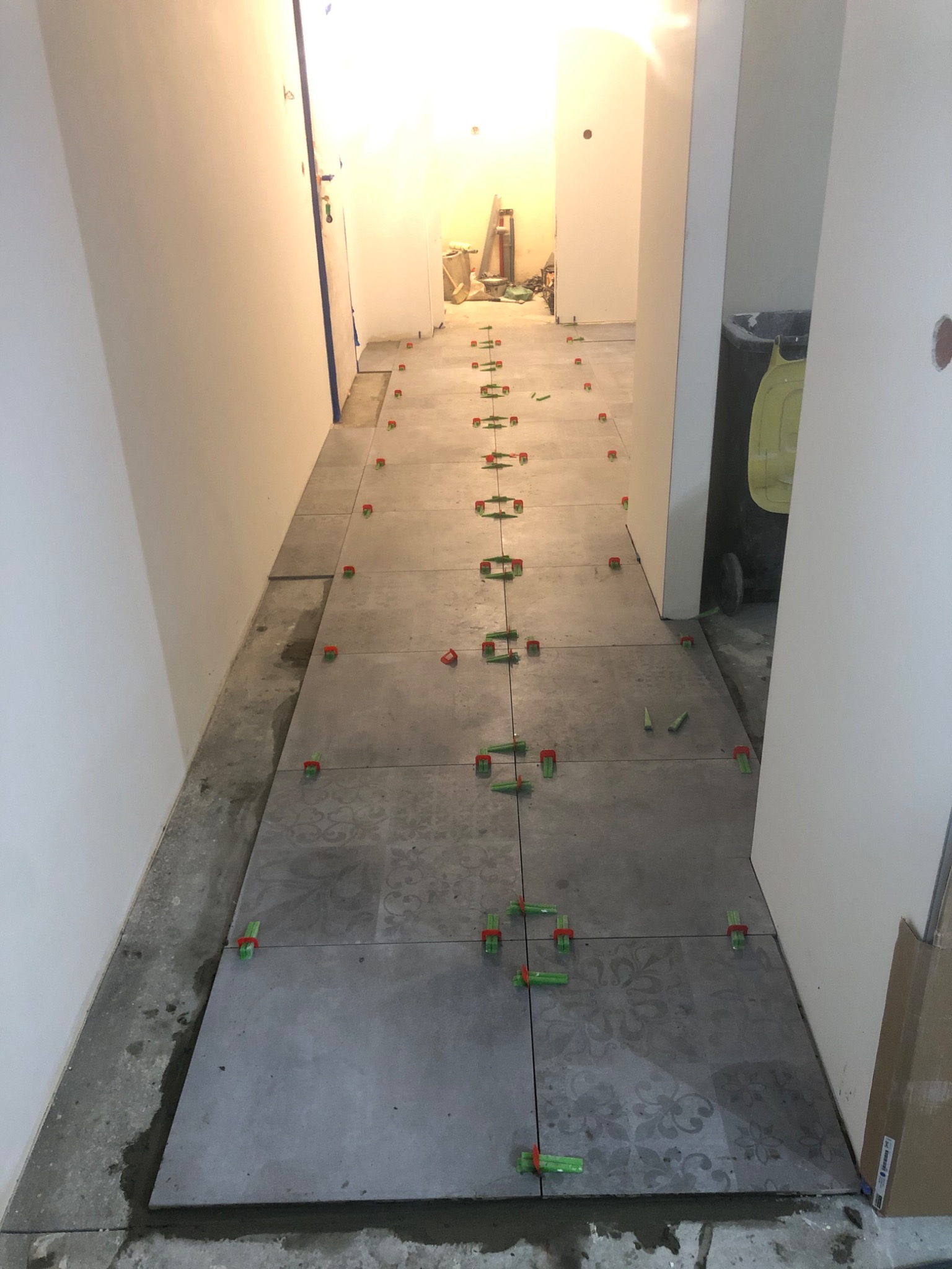 Kładzenie szarych płytek podłogowych z wzorem w korytarzu, z użyciem klipsów poziomujących; widoczne surowe ściany i przygotowane podłoże.