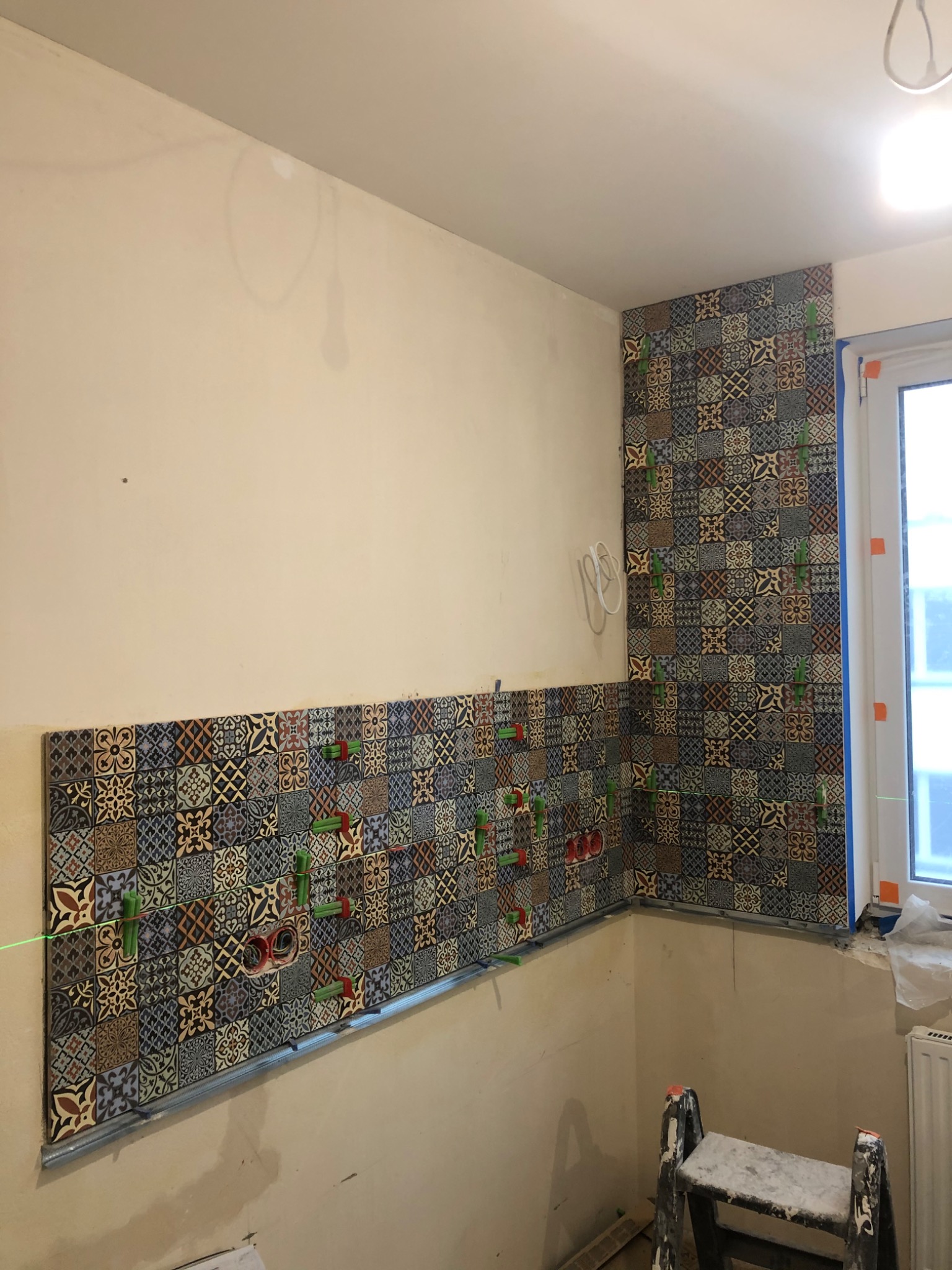 Układanie wzorzystych płytek na ścianie w rogu pomieszczenia, widoczne zielone krzyżyki dystansowe i poziomica laserowa.