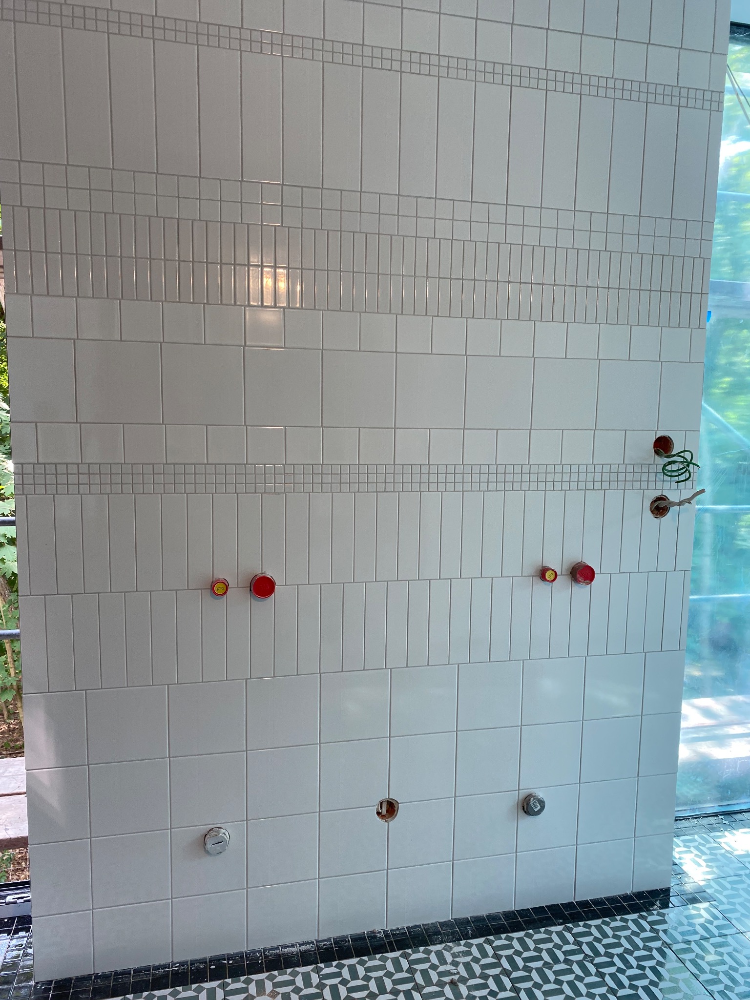 Ściana wyłożona białymi, błyszczącymi płytkami ceramicznymi z dekoracyjnymi pasami z małych kwadratów, widoczne wyprowadzenia instalacji wodnej i elektrycznej oraz fragment podłogi z wzorzystymi...