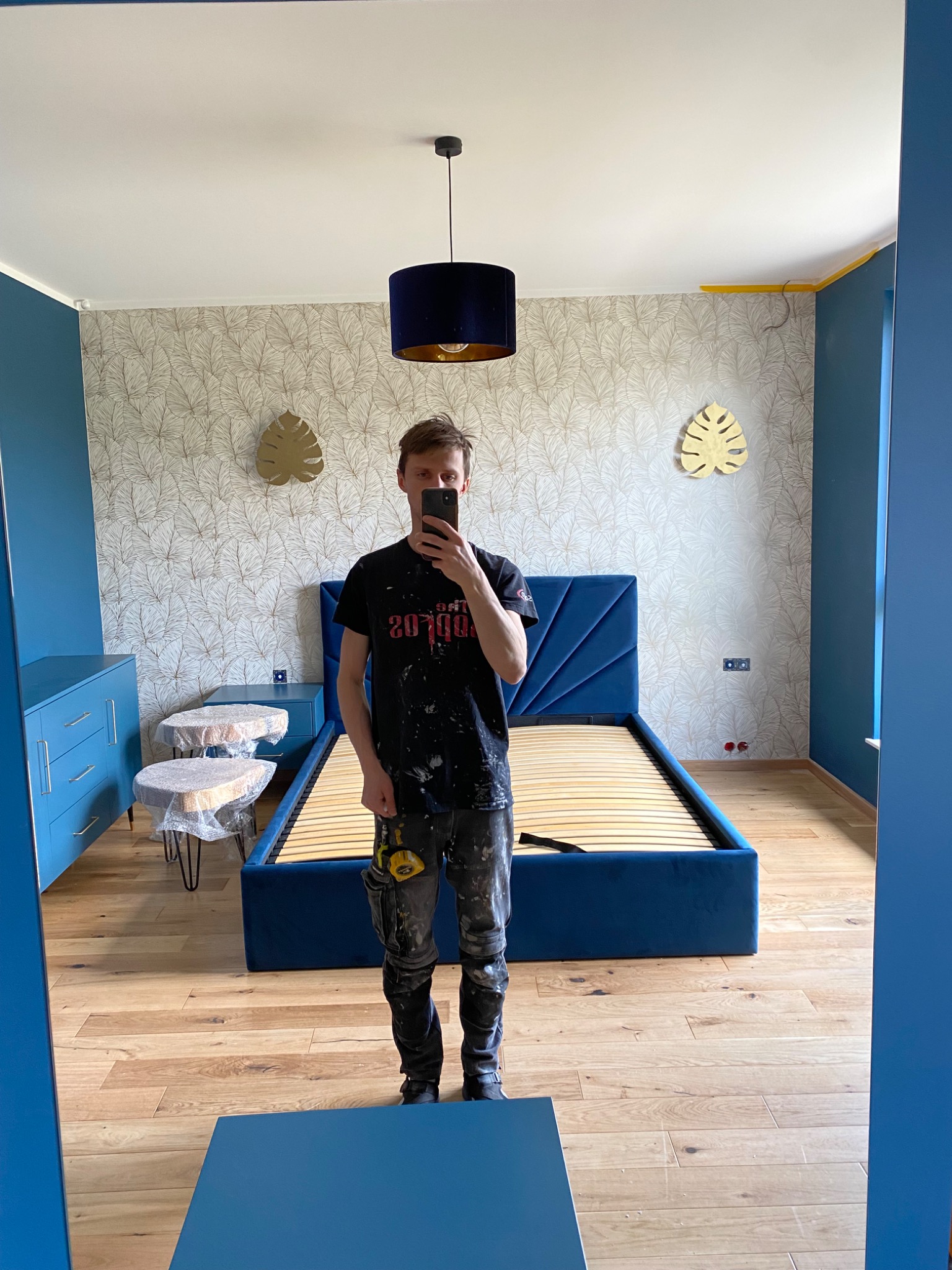 Mężczyzna w ubrudzonym farbą t-shircie robi selfie w lustrze, w tle niebieskie meble, łóżko bez materaca, tapeta w liście i zabezpieczone folią stoliki nocne.