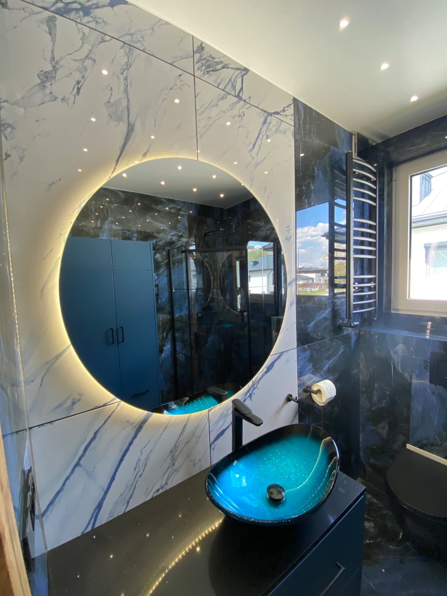 Nowoczesna łazienka z okrągłym lustrem podświetlanym LED, turkusową umywalką na czarnym blacie i marmurowymi płytkami w odcieniach bieli i granatu.