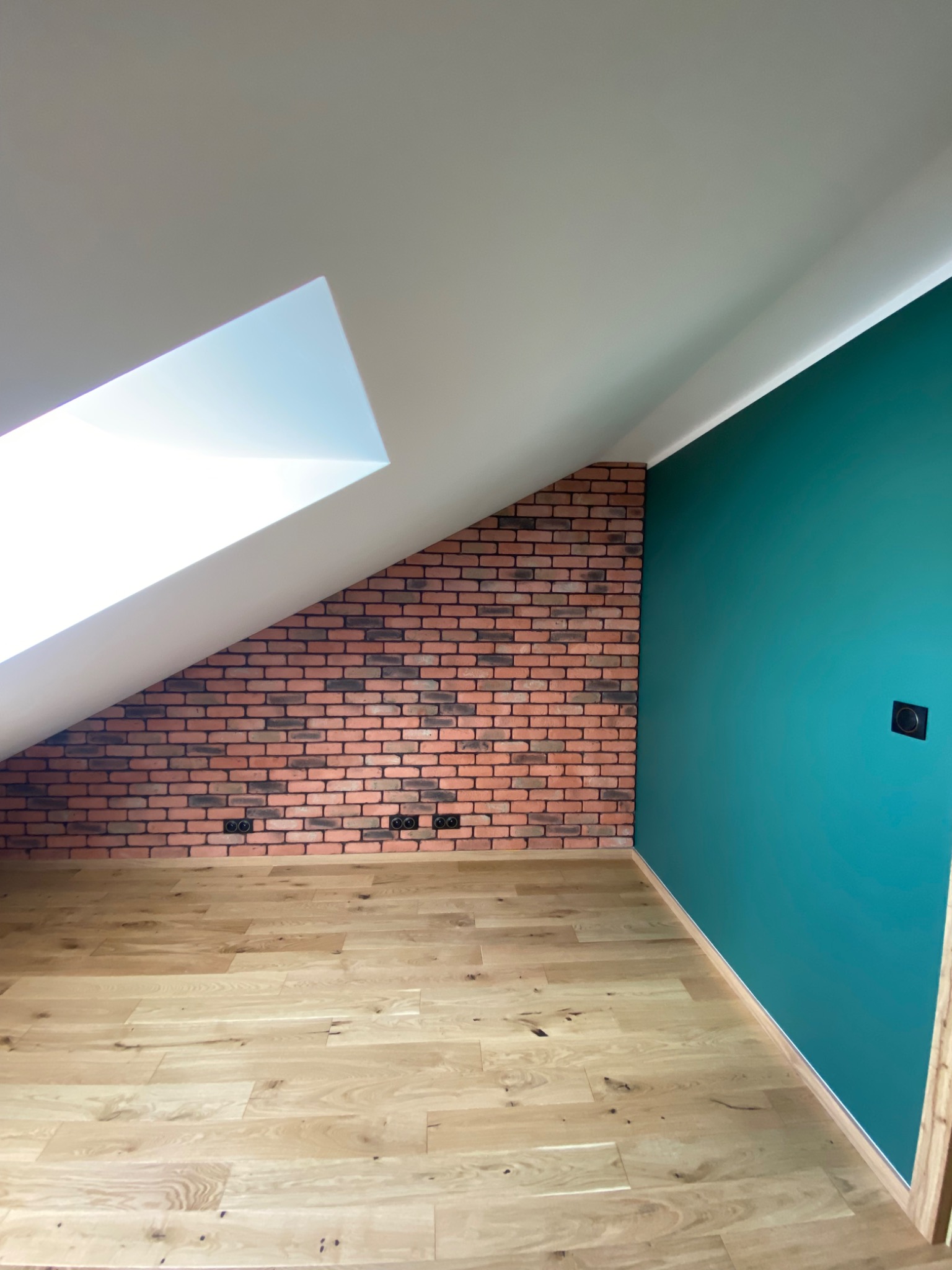 Pokój na poddaszu z drewnianą podłogą, ścianą z cegły klinkierowej pod skosem i turkusową ścianą z gniazdkiem elektrycznym.