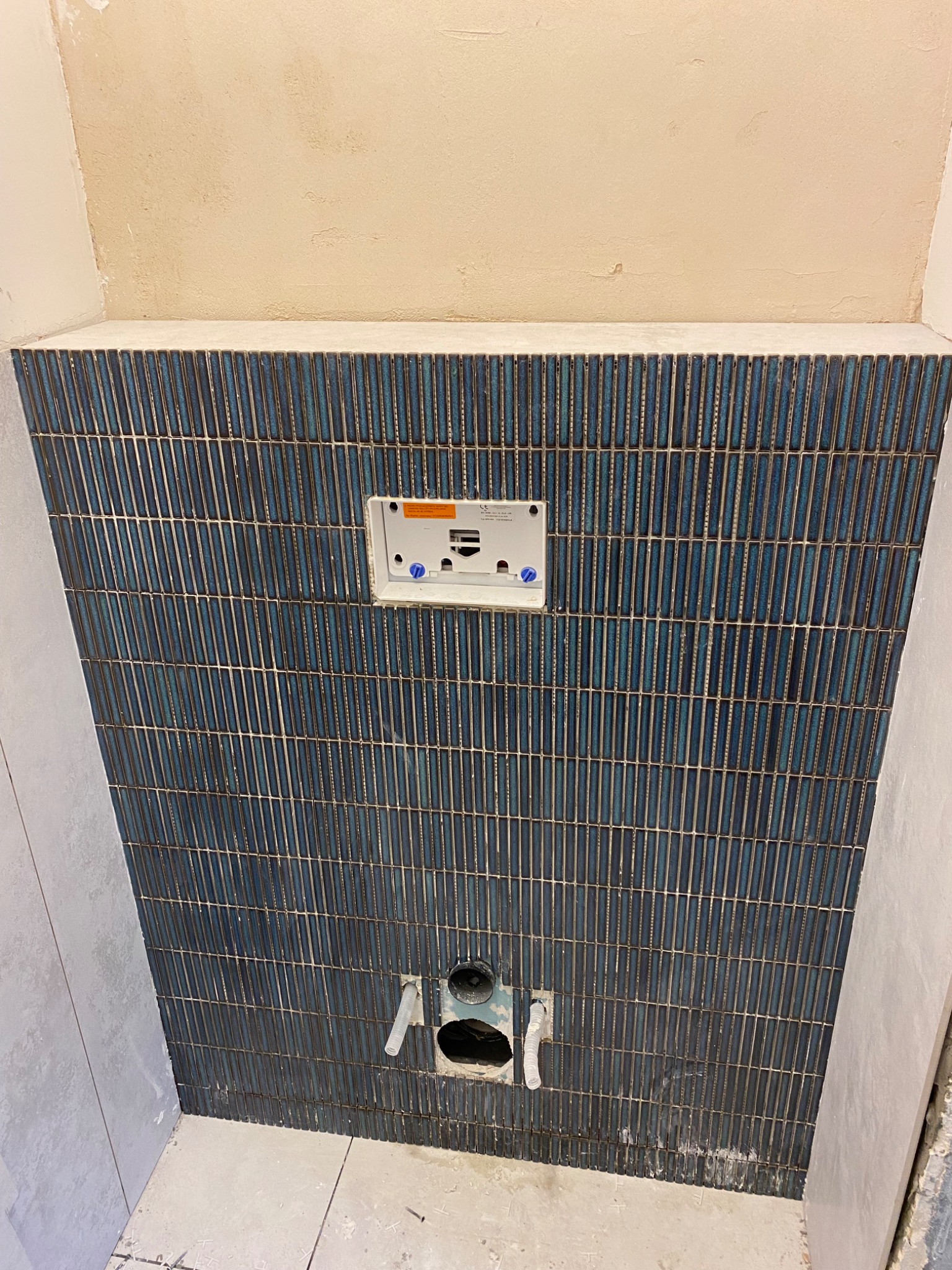 Ściana w łazience wyłożona wąskimi, pionowymi, błyszczącymi płytkami w odcieniach granatu i turkusu, z widoczną instalacją podtynkową i otworami na rury odpływowe, w trakcie prac wykończeniowych.