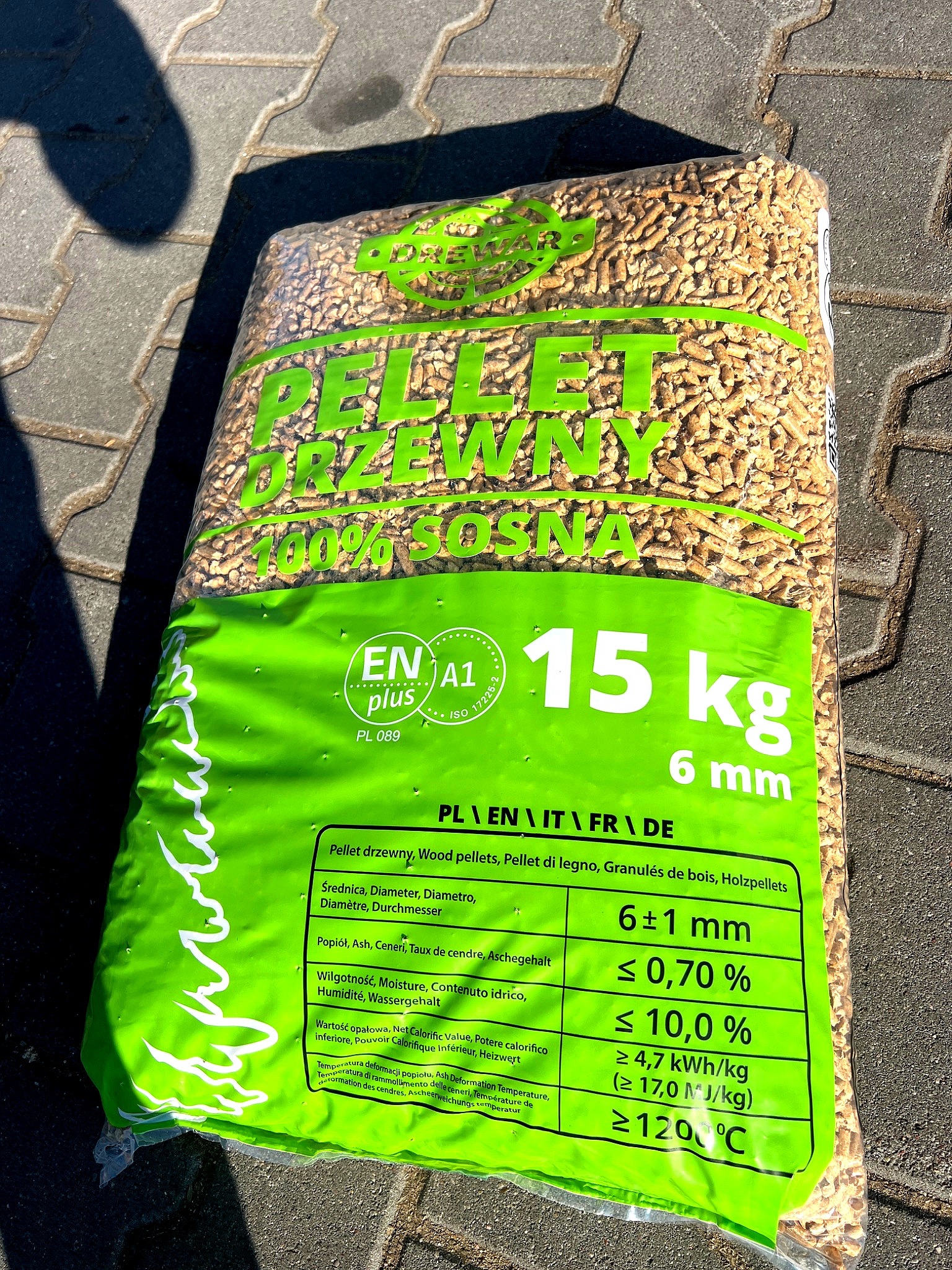 Worek pelletu drzewnego 100% sosna, 15 kg, z certyfikatem ENplus A1, leżący na szarej kostce brukowej. Widoczne parametry techniczne na zielonej etykiecie.