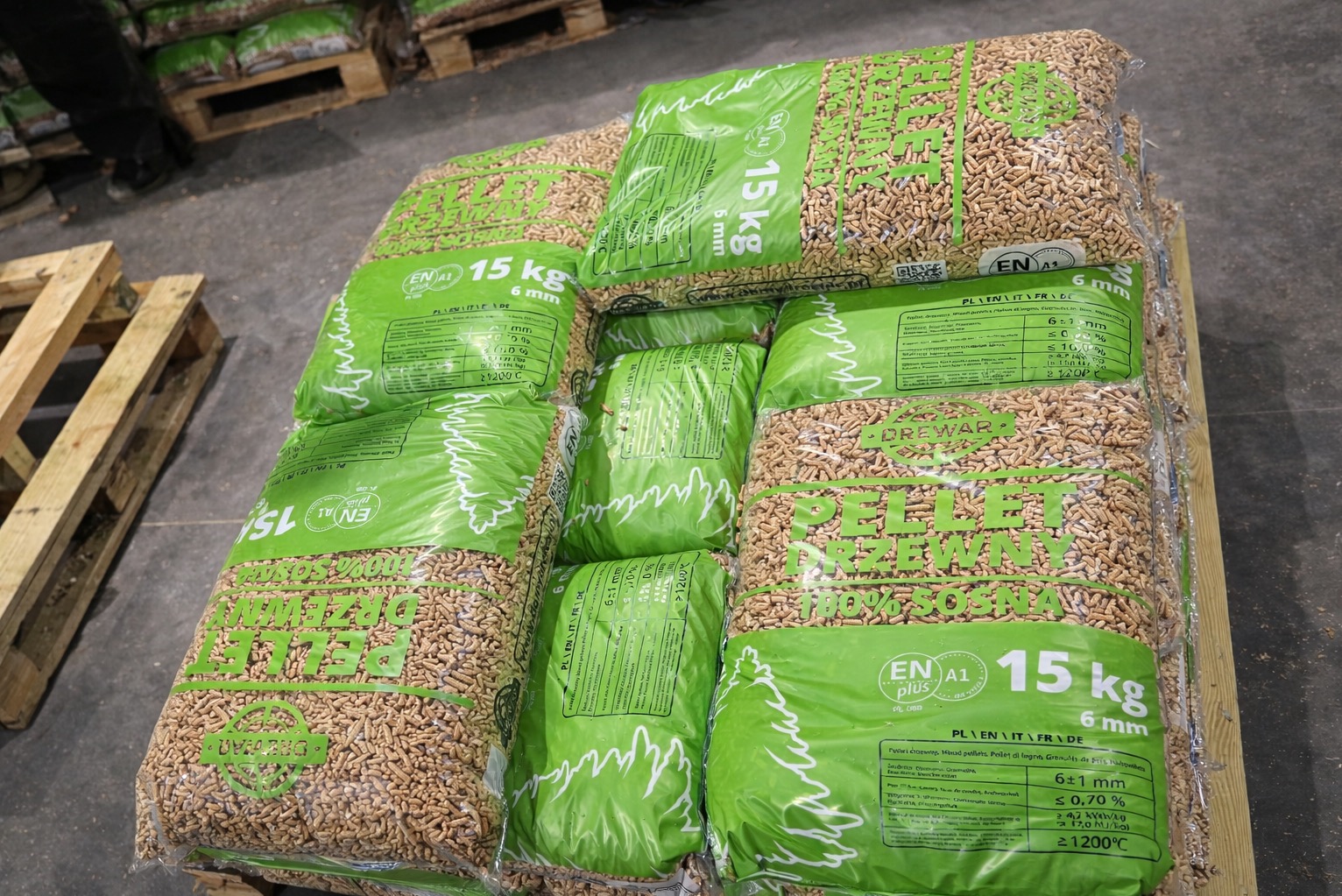 Paleta z workami pelletu drzewnego 15 kg, z widocznym logo producenta i oznaczeniami jakości ENplus A1. Worki w kolorze zielonym, ułożone warstwami na drewnianej palecie.
