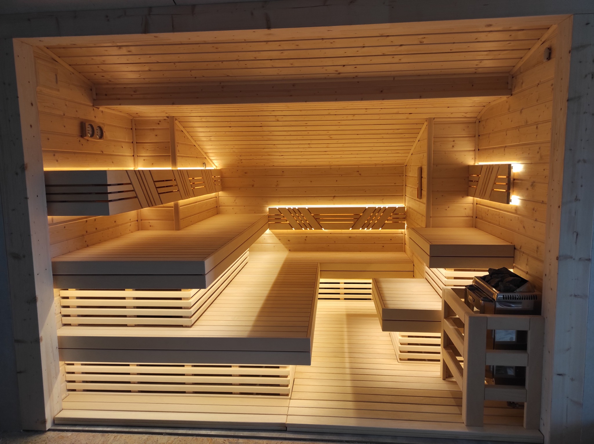 Jasne drewniane wnętrze kabiny z wielopoziomowymi ławkami, podświetleniem LED i piecem na kamienie w rogu.