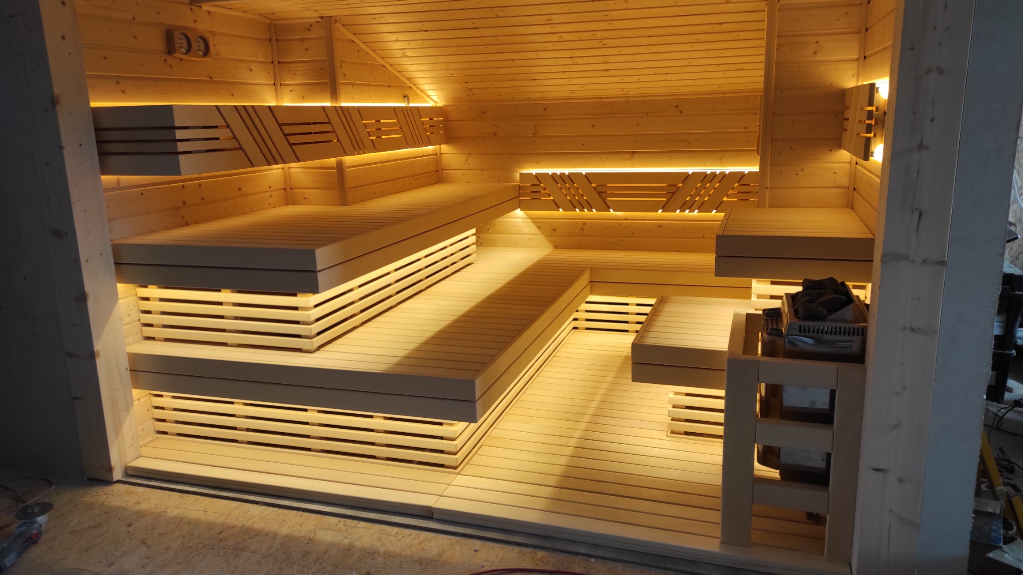 Jasne drewniane wnętrze z podświetlanymi ławkami i piecem z kamieniami, widoczne narzędzia w rogu.