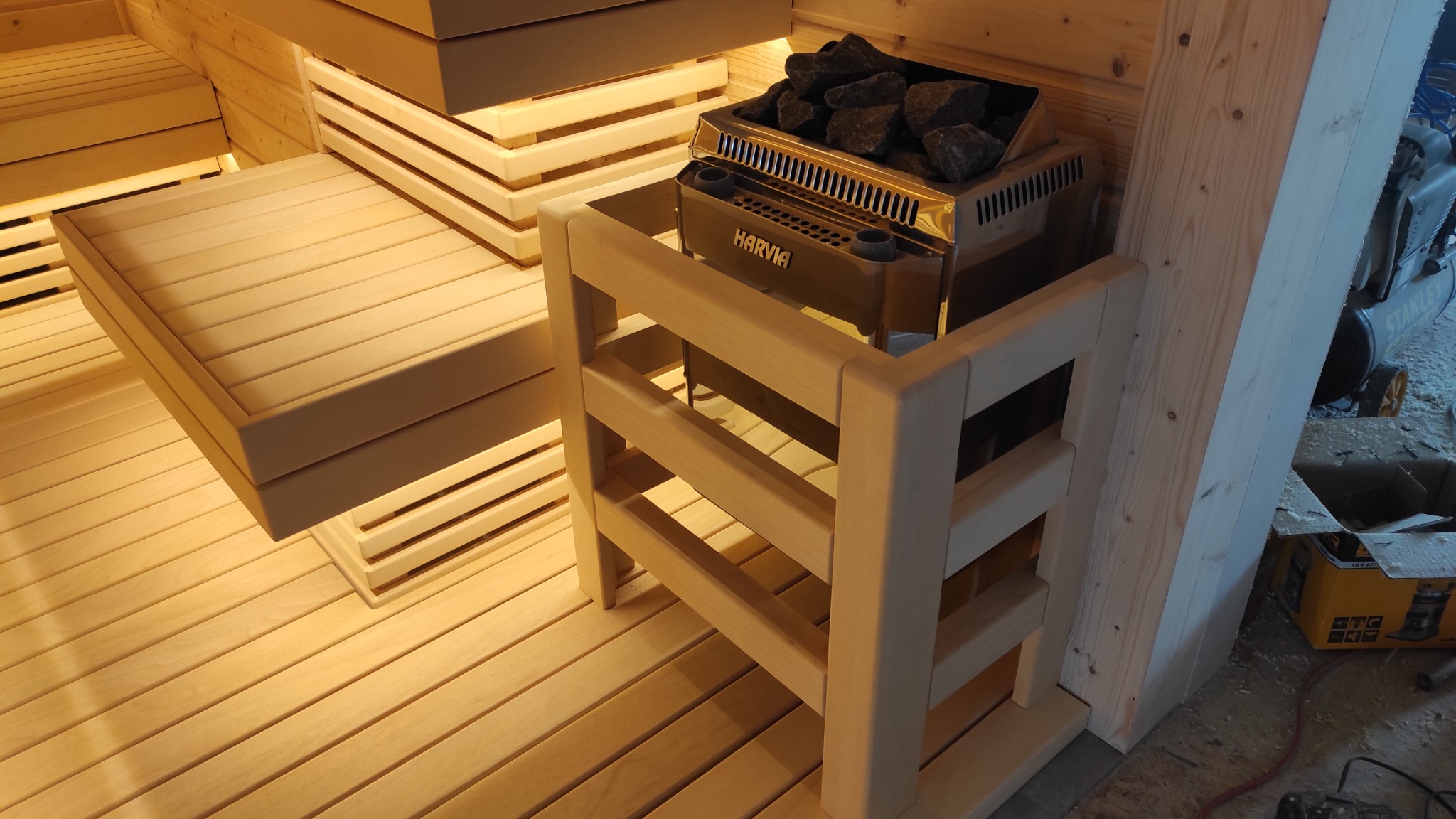 Wnętrze sauny w Myszkowie z drewnianymi ławkami i piecem Harvia z kamieniami, widoczne elementy konstrukcyjne i narzędzia w tle.