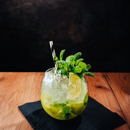 MJS Trade Maciej Śmigiel - Orzeźwiający koktajl mojito z kruszonym lodem, miętą i limonką w szklance na czarnej podkładce, sfotografowany w Topola Mała na drewnianym stole z ciemnym tłem.