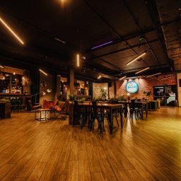 MJS Trade Maciej Śmigiel - Wnętrze przestronnej restauracji z drewnianą podłogą, czarnymi krzesłami i stołami, oświetlone neonowymi lampami i logo marki na ceglanej ścianie.