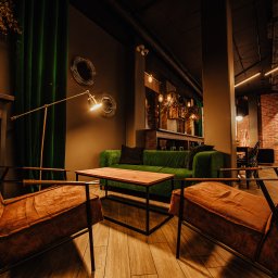 MJS Trade Maciej Śmigiel - Stylowe wnętrze lokalu z dwoma brązowymi fotelami, zieloną sofą i drewnianym stolikiem kawowym, oświetlone lampą podłogową i dekoracyjnymi lampami wiszącymi.