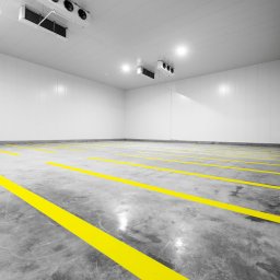 MJS Trade Maciej Śmigiel - Pusta hala chłodnicza z białymi ścianami panelowymi, betonową podłogą z żółtymi pasami wyznaczającymi strefy i klimatyzacją sufitową.