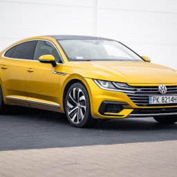 MJS Trade Maciej Śmigiel - Żółty Volkswagen Arteon z czarnym dachem, zaparkowany na kostce brukowej przed białą ścianą, widoczny z boku.