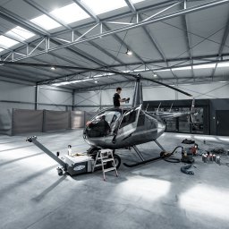 MJS Trade Maciej Śmigiel - Helikopter Robinson R44 Raven II w hangarze podczas prac serwisowych, mechanik na dachu, widoczne narzędzia i sprzęt, oświetlenie sufitowe.