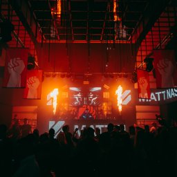 MJS Trade Maciej Śmigiel - DJ Matt Nash występuje na scenie klubu nocnego z efektami pirotechnicznymi, transparentami z symbolem zaciśniętej pięści i tłumem ludzi w tle.