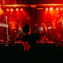 MJS Trade Maciej Śmigiel - DJ Matt Nash w trakcie występu na scenie, otoczony pirotechniką i oświetleniem scenicznym, widoczny z tyłu, z podniesioną ręką w geście entuzjazmu.