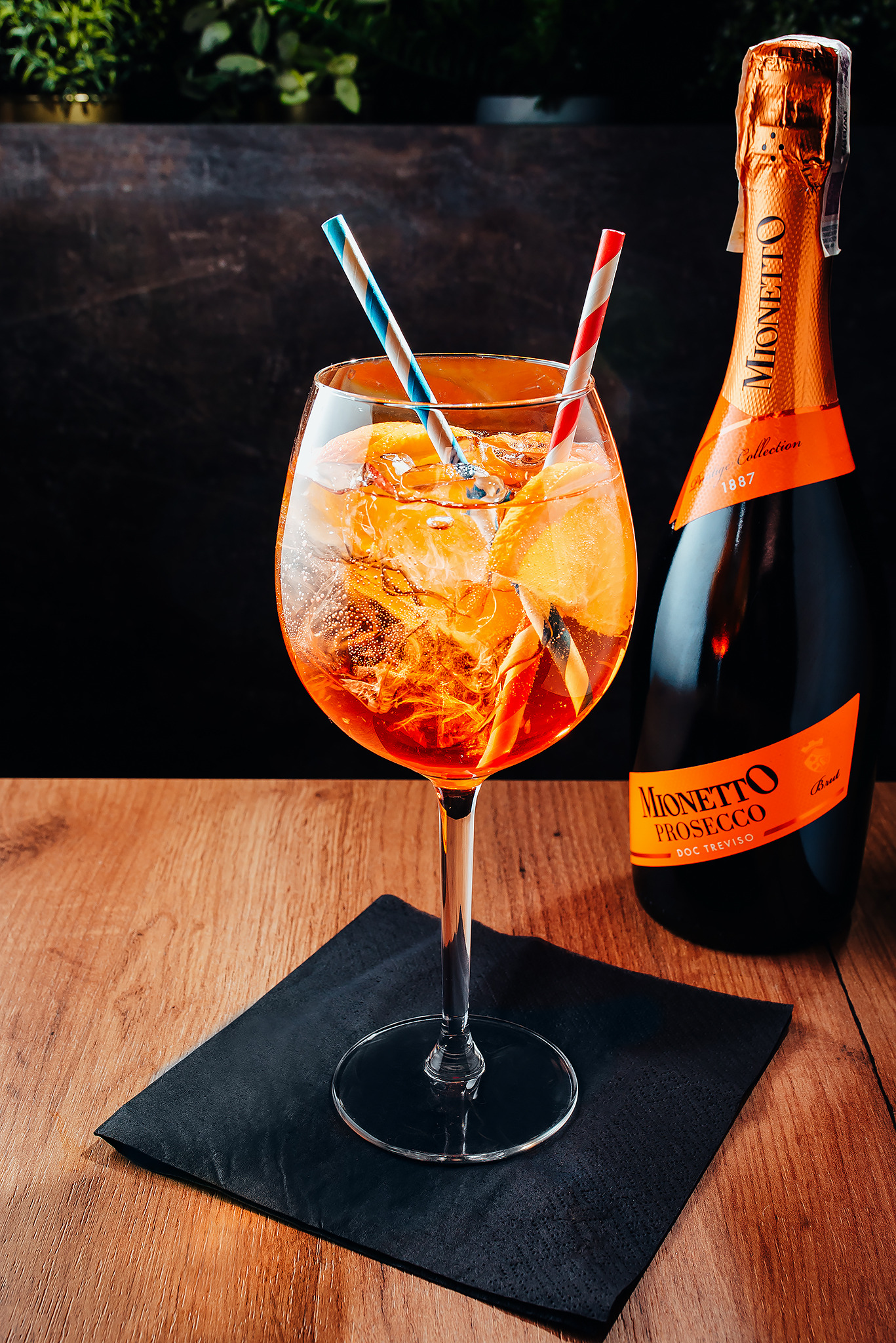 Kieliszek Aperol Spritz z lodem, pomarańczą i kolorowymi słomkami, obok butelka prosecco Mionetto na drewnianym stole z czarną serwetką.