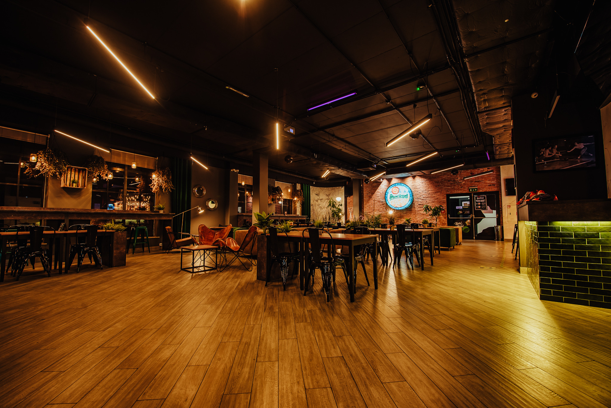 Wnętrze przestronnej restauracji z drewnianą podłogą, czarnymi krzesłami i stołami, oświetlone neonowymi lampami i logo marki na ceglanej ścianie.