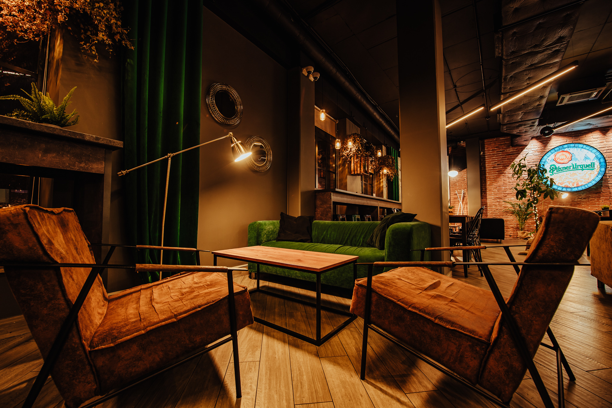 Stylowe wnętrze lokalu z dwoma brązowymi fotelami, zieloną sofą i drewnianym stolikiem kawowym, oświetlone lampą podłogową i dekoracyjnymi lampami wiszącymi.