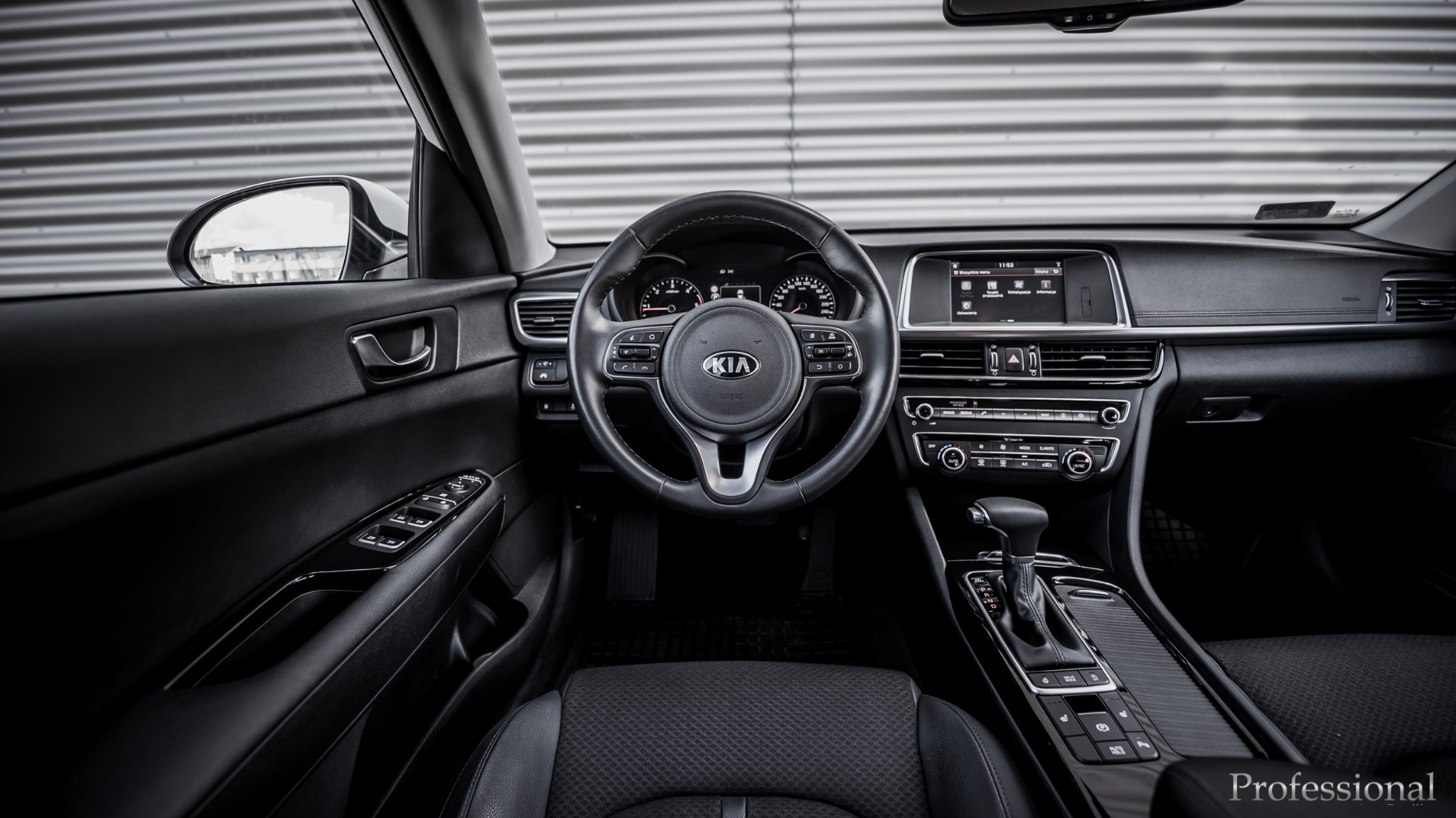 Wnętrze samochodu Kia Optima: deska rozdzielcza, kierownica z logo, panel sterowania klimatyzacją i skrzynia biegów, widok z przodu na tle szarej, metalowej ściany.