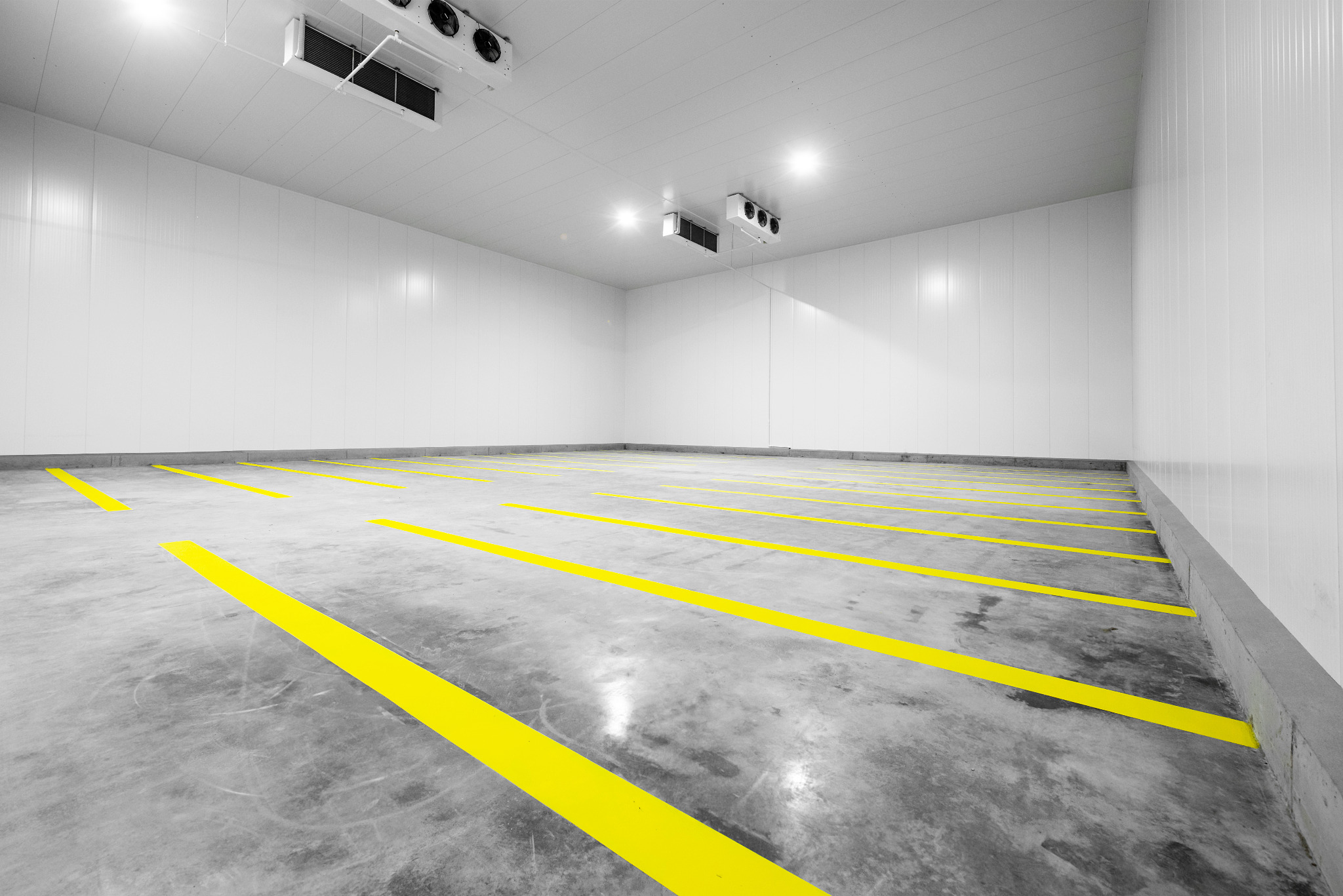 Pusta hala chłodnicza z białymi ścianami panelowymi, betonową podłogą z żółtymi pasami wyznaczającymi strefy i klimatyzacją sufitową.