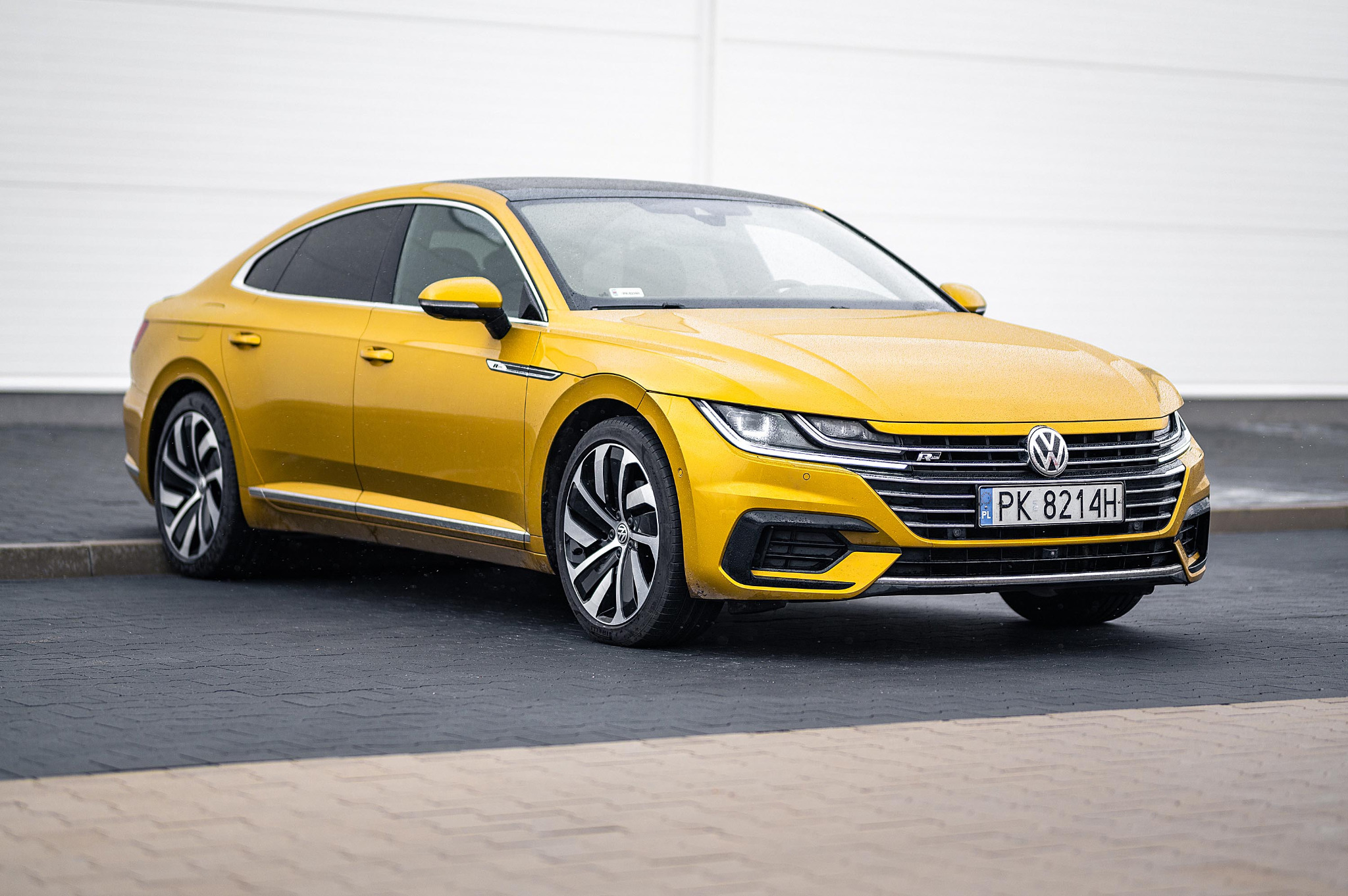 Żółty Volkswagen Arteon z czarnym dachem, zaparkowany na kostce brukowej przed białą ścianą, widoczny z boku.