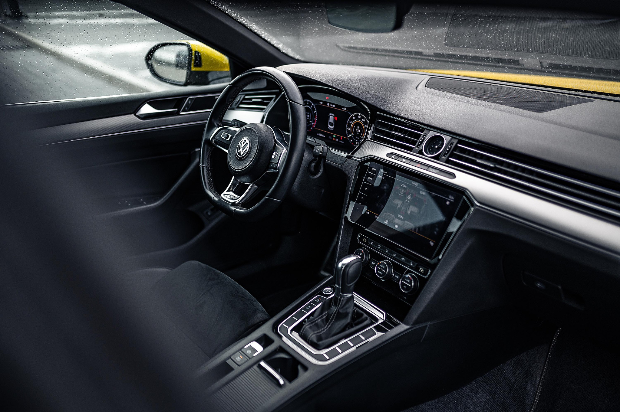 Wnętrze samochodu Volkswagen Passat z czarną deską rozdzielczą, skórzaną kierownicą z logo VW i dotykowym ekranem nawigacji, widoczne krople deszczu na szybie.