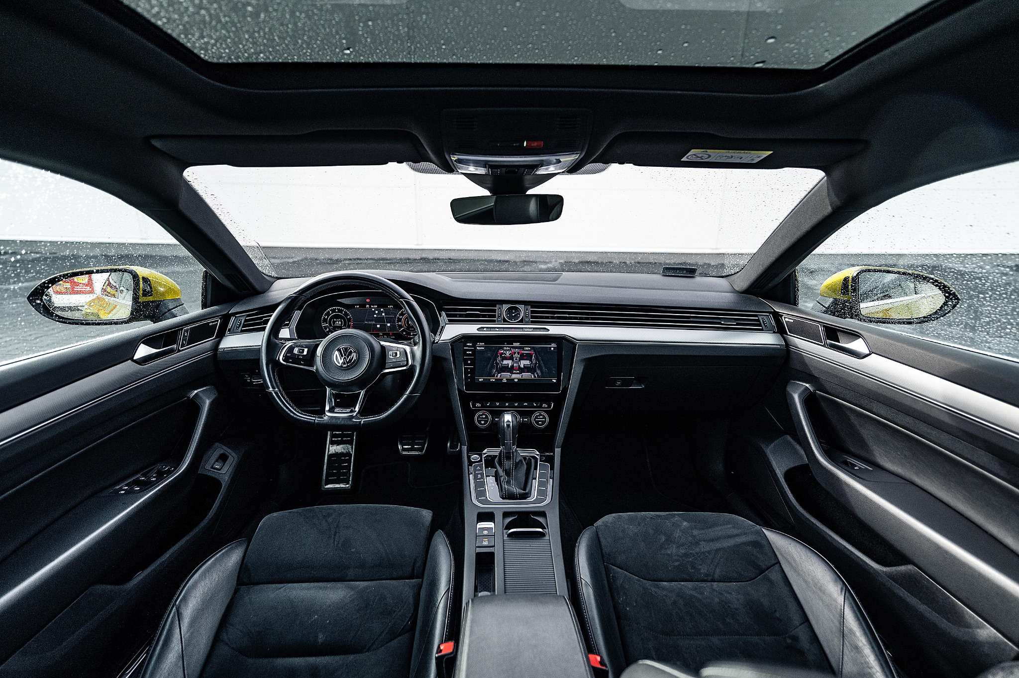 Wnętrze samochodu Volkswagen Arteon z czarną tapicerką, widok z przodu na deskę rozdzielczą, kierownicę i fotele, szyba przednia z kroplami deszczu, widoczny żółty bok innego pojazdu w lusterku.