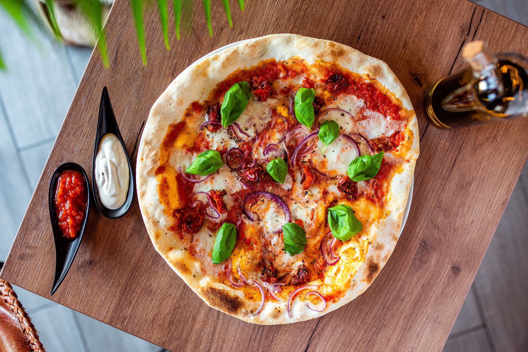 Pizza z pomidorami, czerwoną cebulą i bazylią na drewnianym stole, obok sos pomidorowy i czosnkowy w czarnych miseczkach, butelka oliwy w tle.