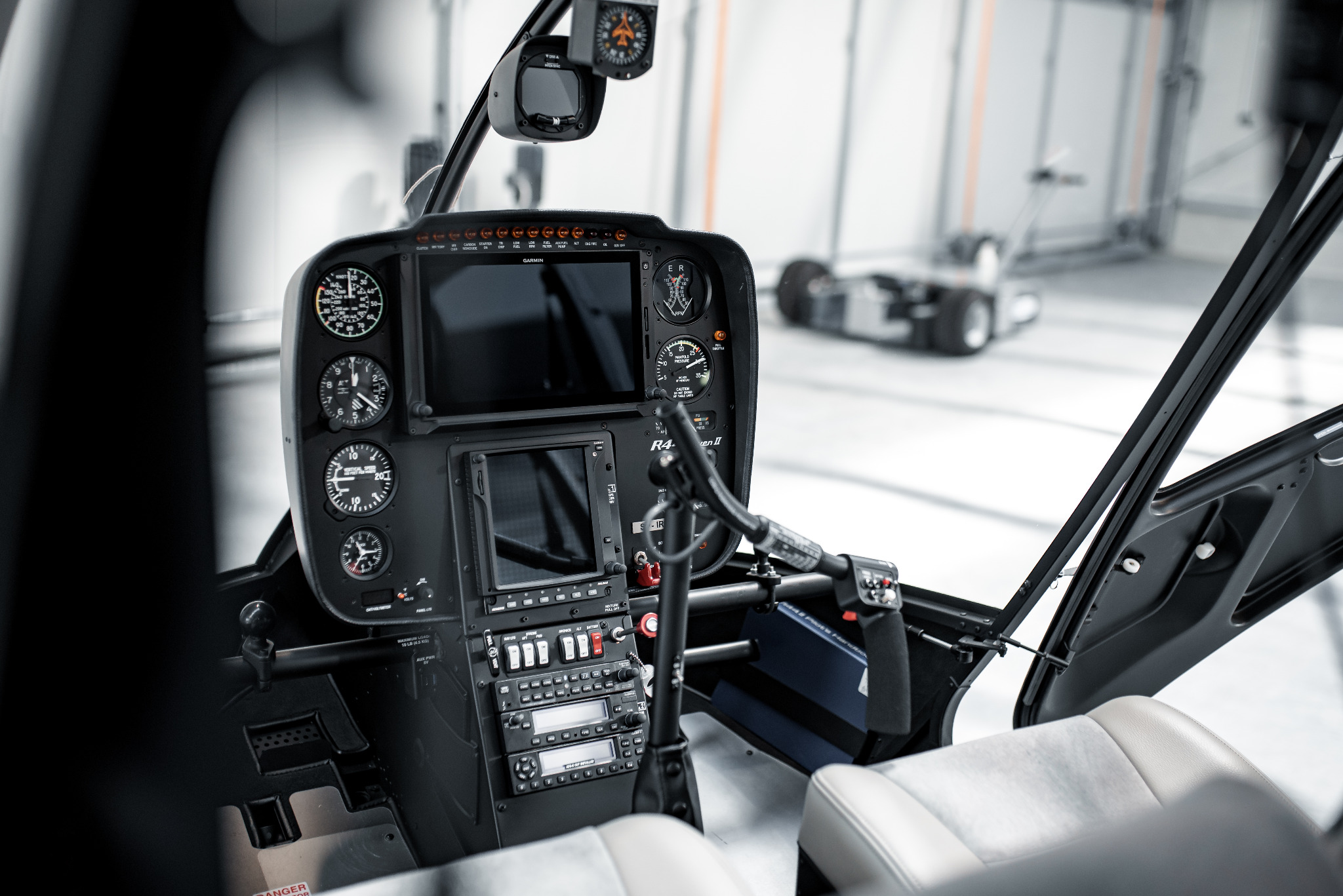 Kokpit helikoptera R44 Raven II z widocznymi wskaźnikami, panelem sterowania i fotelami, sfotografowany w jasnym hangarze z wózkiem transportowym w tle.
