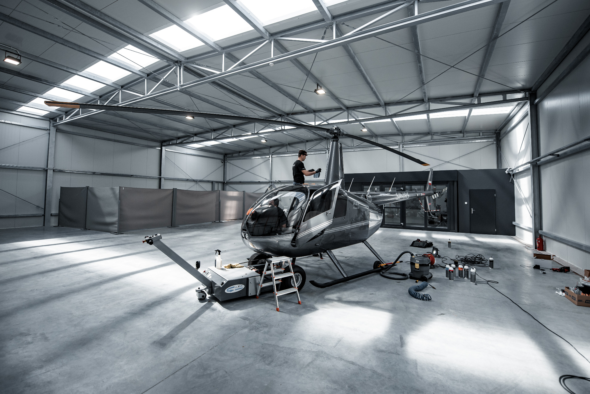 Helikopter Robinson R44 Raven II w hangarze podczas prac serwisowych, mechanik na dachu, widoczne narzędzia i sprzęt, oświetlenie sufitowe.