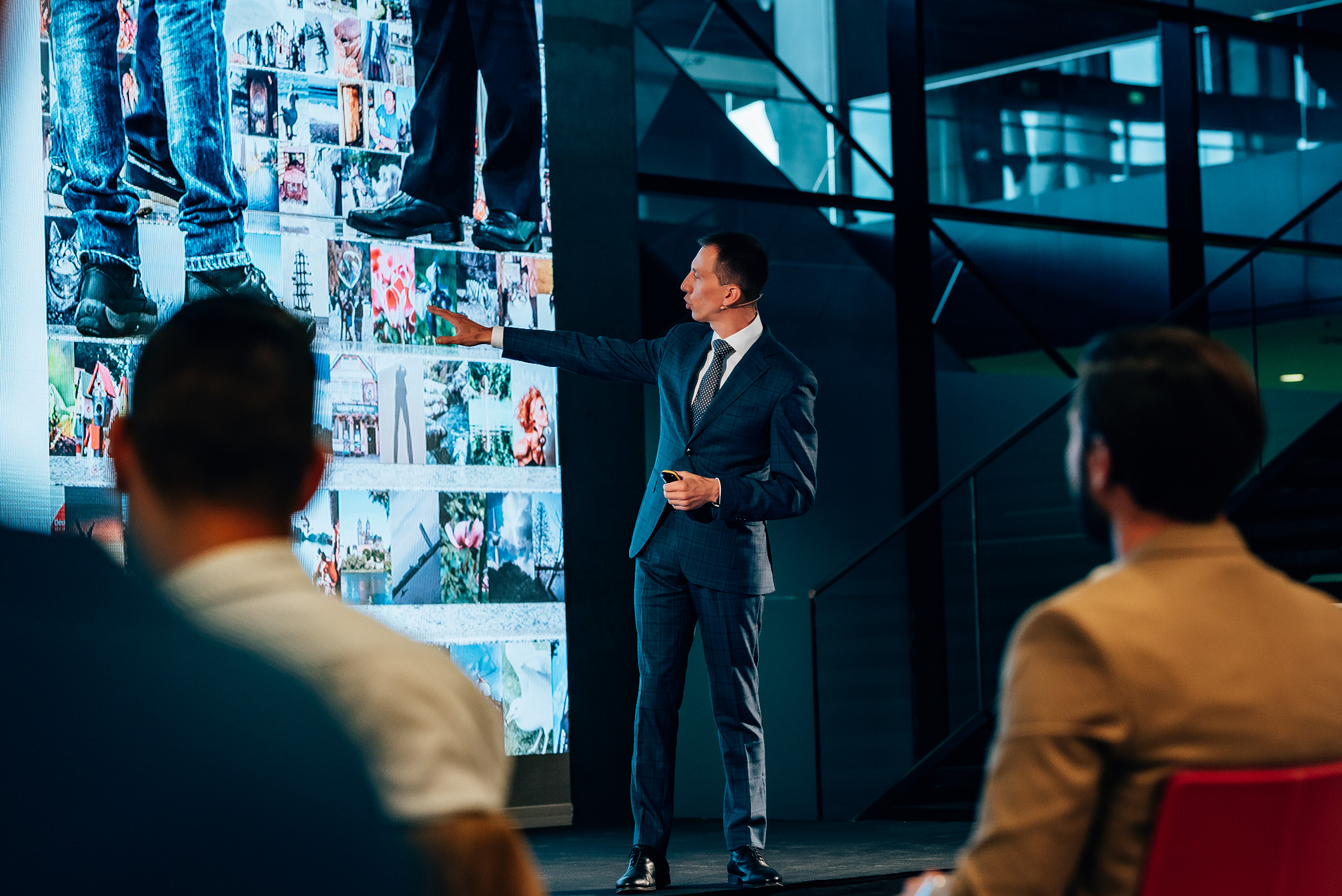 Prelegent w garniturze prezentuje kolaż zdjęć na ekranie podczas konferencji, wskazując palcem na fragment ekspozycji.