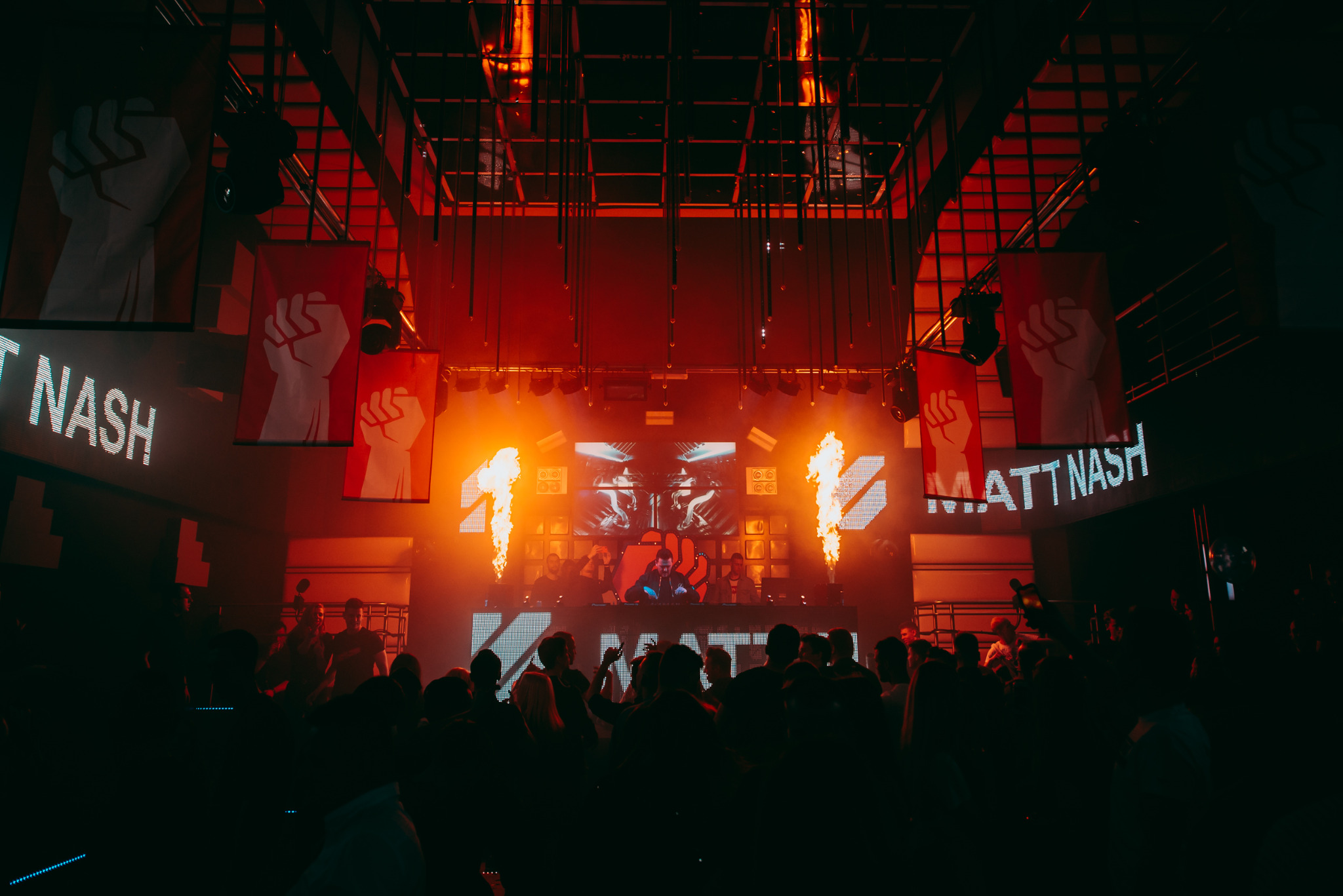 DJ Matt Nash występuje na scenie klubu nocnego z efektami pirotechnicznymi, transparentami z symbolem zaciśniętej pięści i tłumem ludzi w tle.