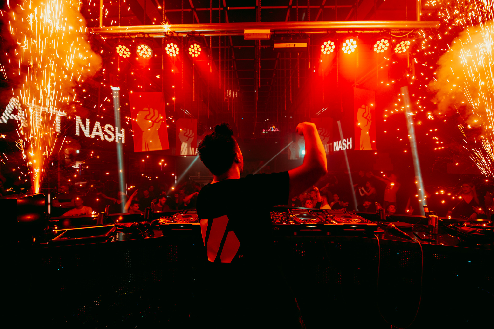 DJ Matt Nash w trakcie występu na scenie, otoczony pirotechniką i oświetleniem scenicznym, widoczny z tyłu, z podniesioną ręką w geście entuzjazmu.