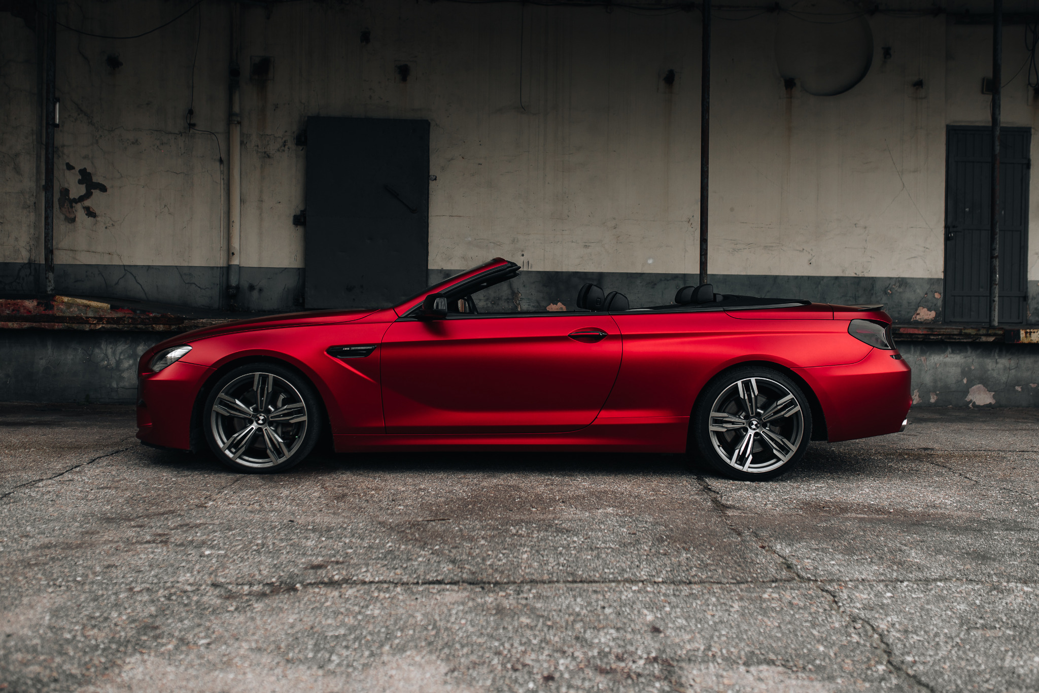 Czerwony kabriolet BMW M6 na tle betonowej ściany z dwiema parami drzwi, z widocznymi śladami użytkowania i obdrapaną farbą.