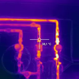 Zdjęcie termowizyjne instalacji grzewczej z widocznymi rurami i elementami pomiarowymi, wskazujące temperaturę 38,1°C, wykonane kamerą FLIR.