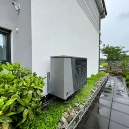 Szara pompa ciepła Alpha Innotec zamontowana na ścianie budynku w Zebrzydowej, otoczona zielenią i kamieniami, widok z boku na tle pochmurnego nieba.