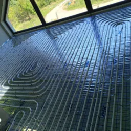 Białe rurki ogrzewania podłogowego ułożone w pętle na niebieskiej folii z nadrukiem kratki, widok z g&oacute;ry, okno w tle.