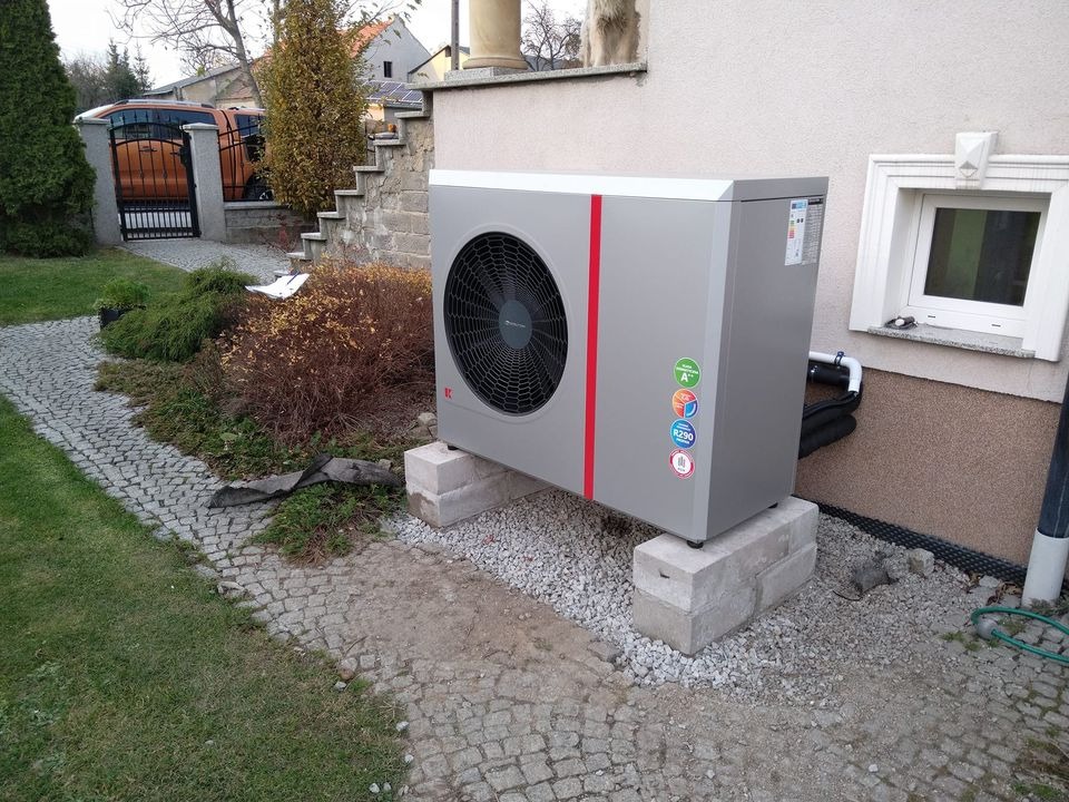 Szara jednostka zewnętrzna z czerwoną linią, wentylatorem i naklejkami informacyjnymi, umieszczona na betonowych bloczkach przy ścianie domu z małym oknem piwnicznym, widoczne rury i kabel, kostka...