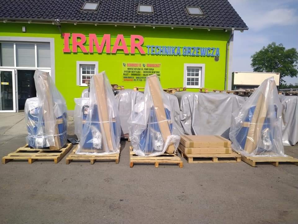 Nowe urządzenia grzewcze, zapakowane w folię, stoją na paletach przed zielonym budynkiem firmy IRMAR Technika Grzewcza.