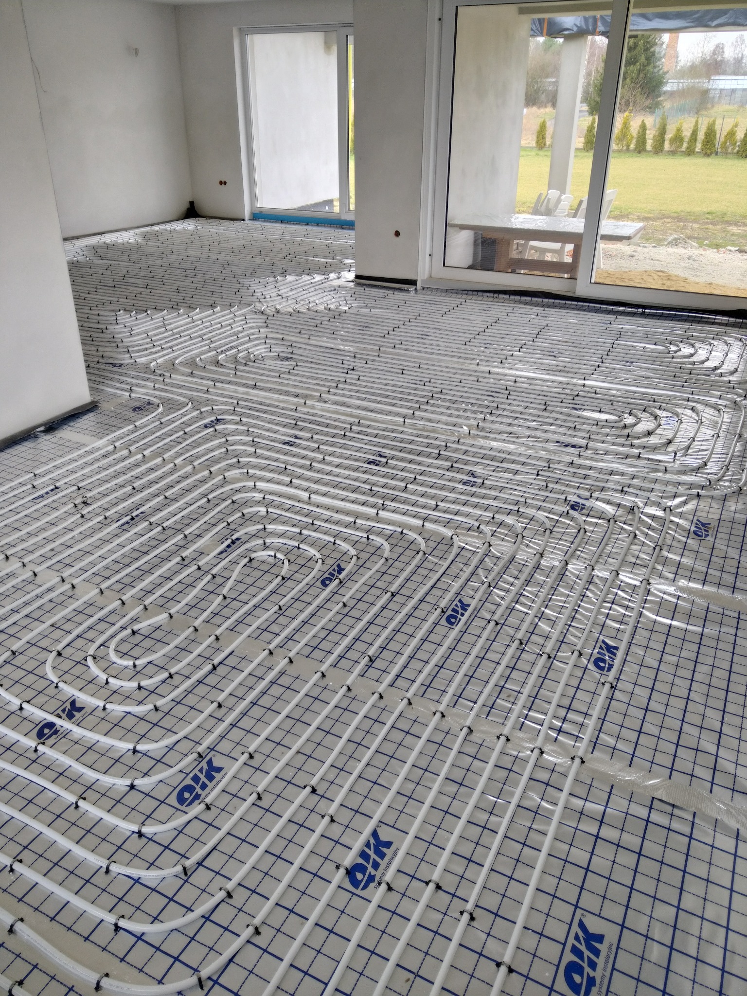 Białe rurki ogrzewania podłogowego ułożone w pętle na folii z nadrukowaną siatką, widoczne duże okna w surowym stanie.