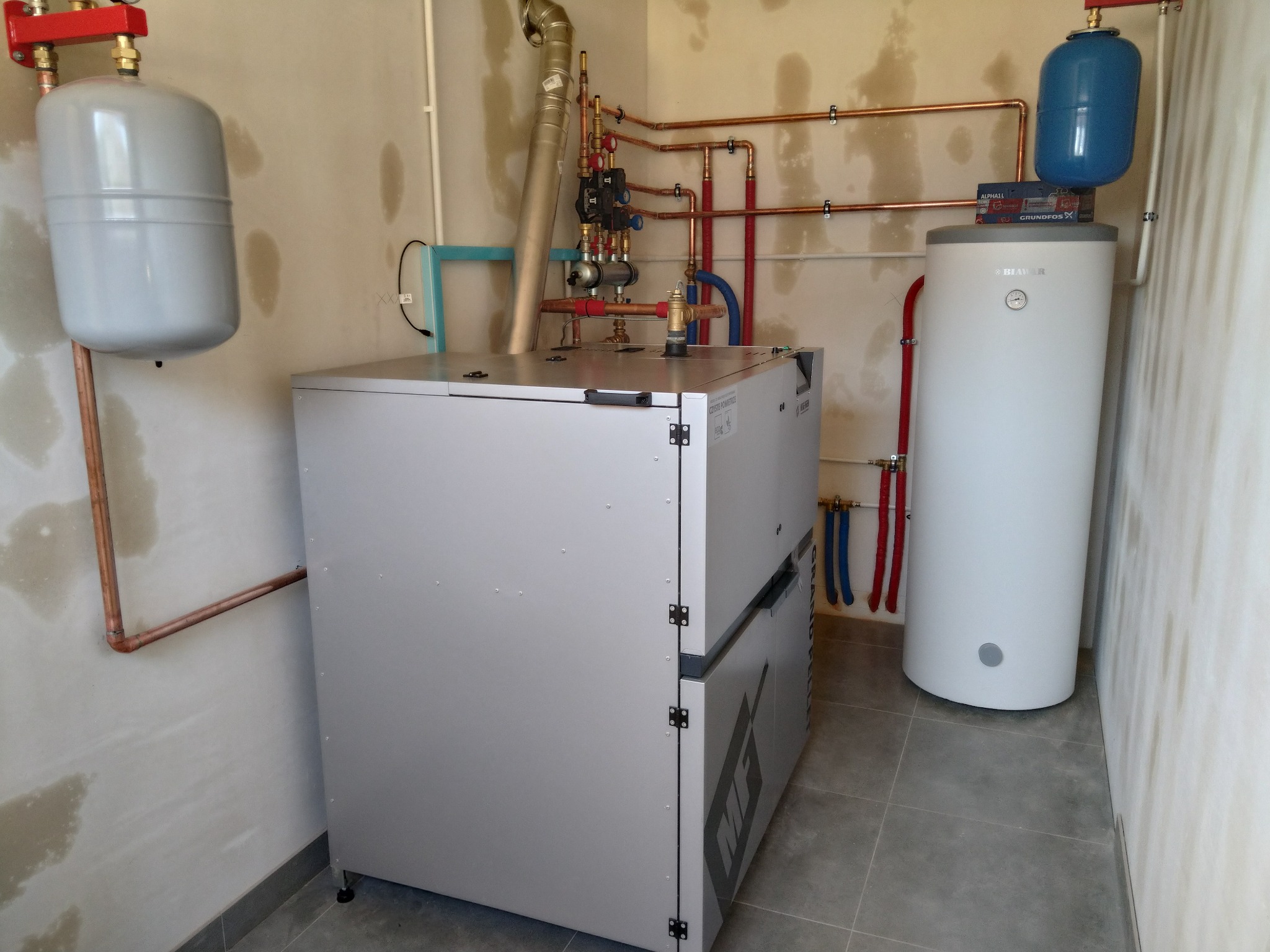 Wnętrze kotłowni z zainstalowanym piecem na pellet, zbiornikiem akumulacyjnym i rozbudowaną instalacją hydrauliczną z rur miedzianych, w surowym wykończeniu ścian.