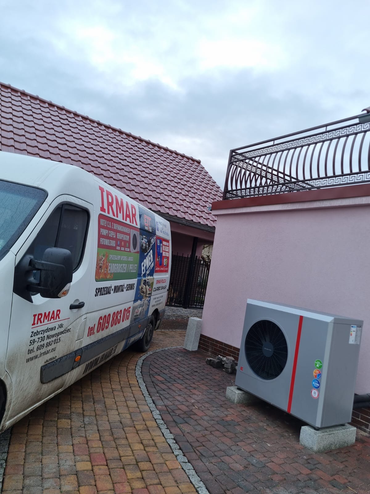 Szara pompa ciepła z czerwoną linią, zamontowana na betonowych bloczkach obok różowego budynku z ozdobnym balkonem, na kostce brukowej, z widocznym brudnym samochodem dostawczym firmy IRMAR.