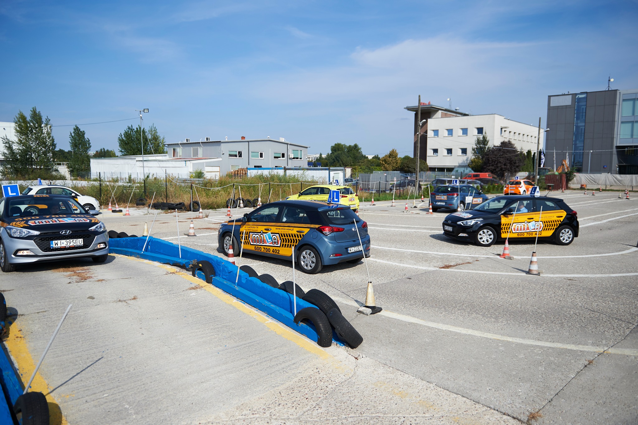 Plac manewrowy z samochodami oznaczonymi literą L, przygotowanymi do nauki jazdy, otoczony pachołkami i elementami toru przeszkód, w tle budynki.