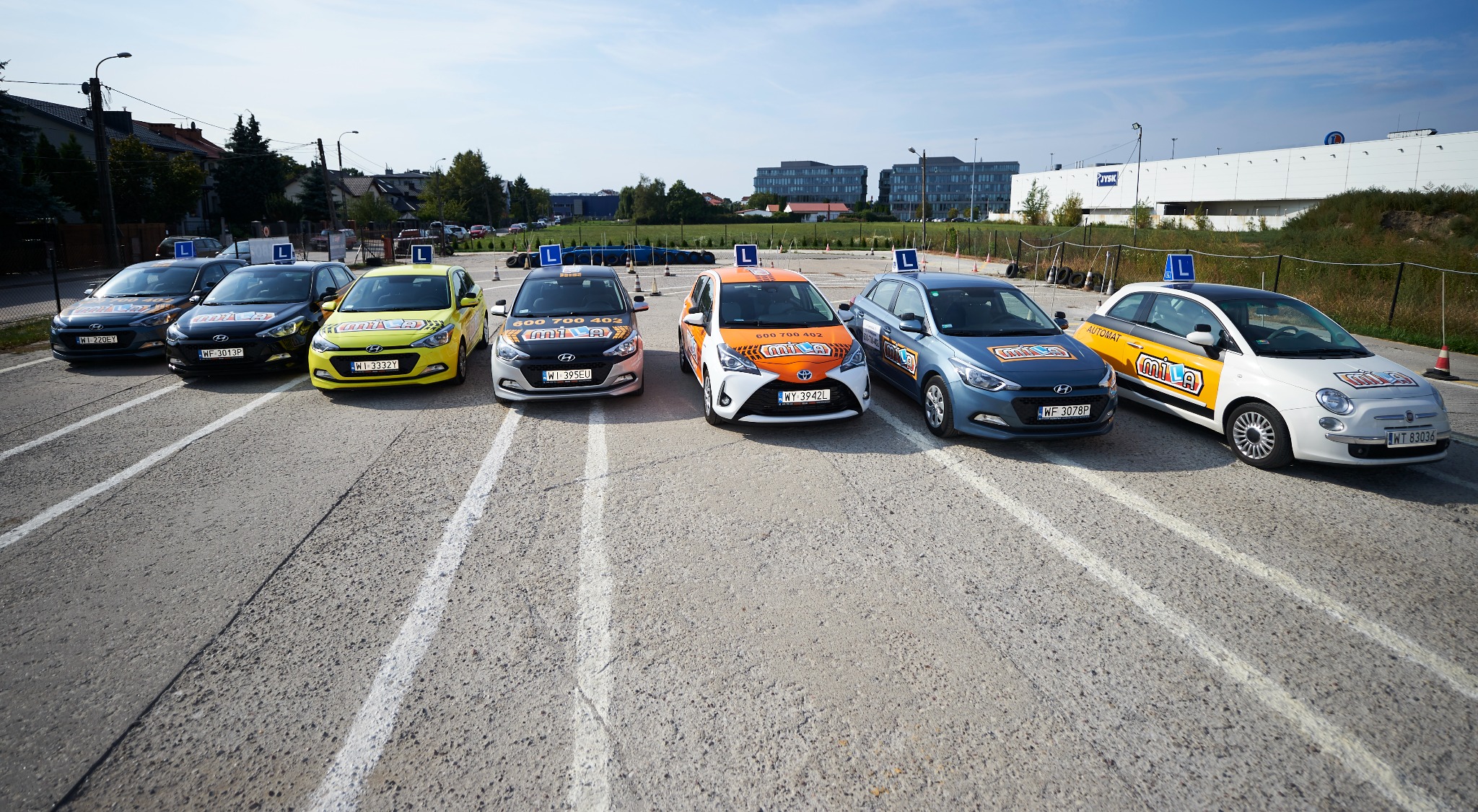 Flota samochodów o różnych kolorach, oznaczonych literą 'L', zaparkowana na placu manewrowym w Warszawie. Widoczny budynek JYSK w tle.